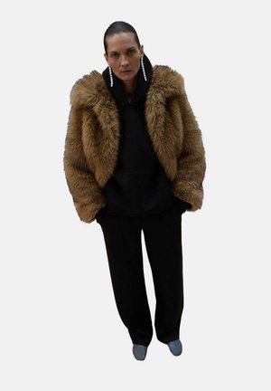 Veste en fausse fourrure marron sur un hoodie noir, associée à un pantalon noir et des chaussettes bleu clair. Elle possède un grand col et des boucles d'oreilles pendantes.