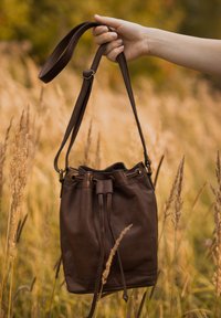 Borsa a secchiello in pelle marrone con tracolla regolabile, chiusura a coulisse e texture goffrata, tenuta davanti a uno sfondo di erba alta.