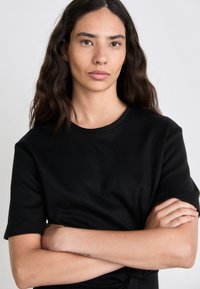 T-shirt in cotone nero con maniche corte, scollo rotondo e texture morbida. Il modello ha le braccia incrociate, mostrando un design minimalista.