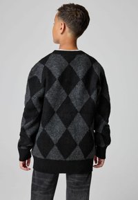 Ragazzo con capelli corti che indossa un maglione a motivo diamante nero e grigio, girato di spalle su uno sfondo grigio semplice.