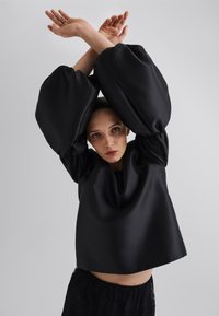 Svart oversized topp med store, rynkede ermer. Glatt, skinnende tekstur, rund halslinning og løs passform. Kombinert med svarte blonderbukser.