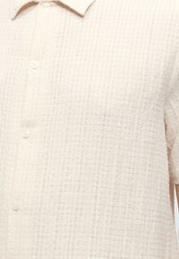 Gros plan sur une chemise beige texturée à manches courtes avec col et boutons blancs sur le devant.