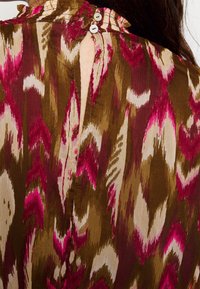 Pestrý top s hnědým pozadím a růžovými a krémovými vzory, volánkovým výstřihem a knoflíkovým zapínáním na zadní straně.
