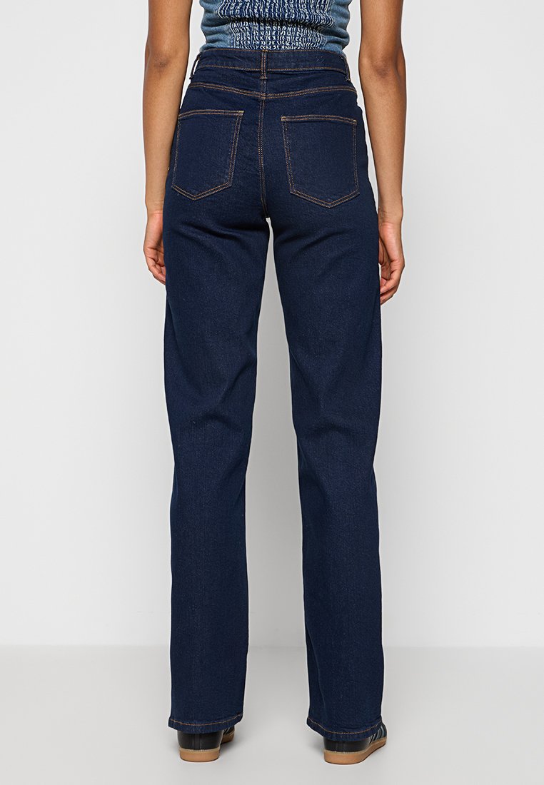 Hoch sitzende dunkelblaue Jeans mit einer lockeren Passform, ausgestattet mit zwei Gesäßtaschen und kontrastierenden Nähten, kombiniert mit schwarzem Schuhwerk.