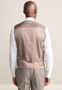 Back view of a light brown satin vest with a tie at the back, worn over a white shirt, paired with checkered grey trousers.