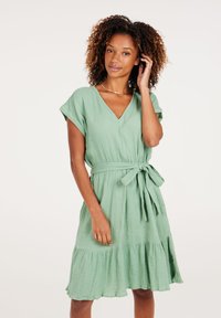 Grünes, kurzärmliges Kleid mit V-Ausschnitt, tailliertem Bund und bindbarem Detail. Verfügt über einen gestuften Rüschensaum und texturierten Stoff.