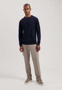Marineblauer Rundhals-Pullover aus Strickstoff, eng anliegende lange Ärmel. Kombiniert mit beigefarbenen geraden Hosen und grauen Schnürschuhen.