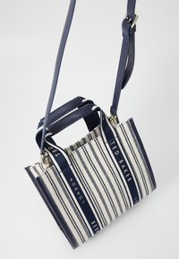 Sac fourre-tout rayé bleu marine et crème avec doubles poignées, bandoulière ajustable et accents de logo. Présente un tissu texturé et une fermeture éclair.