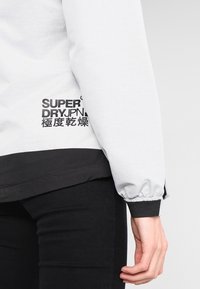 Nærbillede af en person iført en lysegrå jakke med "SUPER DRY JPN" og japansk tekst på ryggen, parret med sorte bukser.