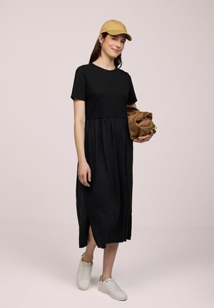 Mujer con vestido midi negro y gorra beige, llevando un bolso marrón, caminando con zapatillas blancas sobre un fondo liso.