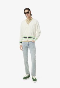 Cardigan bianco a maglia con lavorazione a trecce, con strisce verdi e blu navy sull'orlo, abbinato a un polo beige e jeans azzurri chiari. Scarpe da ginnastica verdi completano il look.