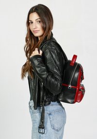 Mochila de imitación de cuero negra y roja con dos cremalleras, correas ajustables y un acabado brillante, llevada en el hombro. Tejido texturizado en la chaqueta.