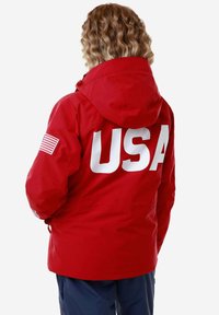 Felpa impermeabile rossa con una grande scritta bianca "USA" sul retro. Presenta un cappuccio e un'adesivo della bandiera americana sulla manica. Tessuto liscio e resistente.