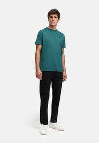Teal kortermet t-skjorte med rund hals og lite logo på venstre bryst, sammen med sorte bukser og hvite sneakers.