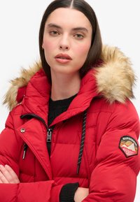 Chaqueta acolchada roja con capucha de pelo sintético, frontal con cremallera y cordones negros. Presenta un parche con el logo en la manga y un diseño acolchado.