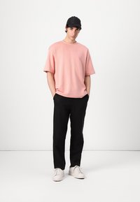 Ροζ κοντομάνικο μπλουζάκι, μαύρο παντελόνι και λευκά αθλητικά παπούτσια. Το μοντέλο στέκεται με τα χέρια στις τσέπες, φορώντας μαύρο καπέλο. Απλός, καθαρός σχεδιασμός.