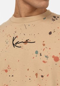 Béžové bavlněné tričko s flekovaným vzorem v červené, modré a hnědé barvě. Černě vyšitý podpis na hrudi. Kulatý výstřih.