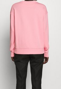 Personne portant un sweat-shirt rose ample et un pantalon noir, vue de dos sur un fond uni.