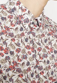 Camisa floral con base blanca, que presenta flores rojas y azules, hojas verdes, un cuello con botones y una textura suave y lisa.