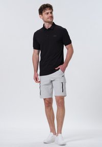 Sort polo-shirt med et diskret logo, parret med lysegrå cargo-shorts med sidelommer og sorte lynlåse, kompletteret med hvide sneakers.