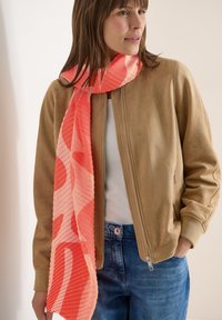 Femme portant une veste zippée beige, un haut blanc, un jean bleu, et une longue écharpe rouge à motifs drapée autour du cou.