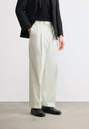 Osoba ubrana w białe spodnie o szerokich nogawkach, czarną marynarkę, czarną koszulę i czarne eleganckie buty, stojąca na szarej podłodze na tle białej ściany.