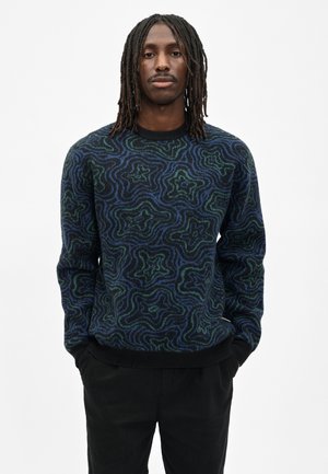 Man met dreadlocks tot op de schouders, die een blauwgroen gebreide trui en zwarte broek draagt, handen in de zakken.