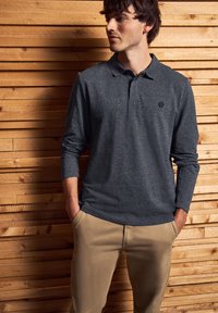 Muž v šedém polo tričku s dlouhými rukávy a béžových kalhotách, stojící s rukama v kapsách u dřevěné stěny.