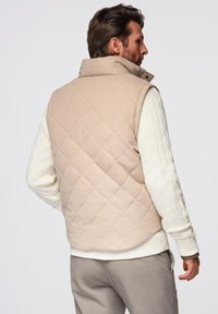 Vaalean beige hitsatun liivin, jossa on korkea kaulus, sileä pinta ja hieman pyöristetty helma, päällä valkoinen puuvillaneule, jossa on kaapelikudonta.