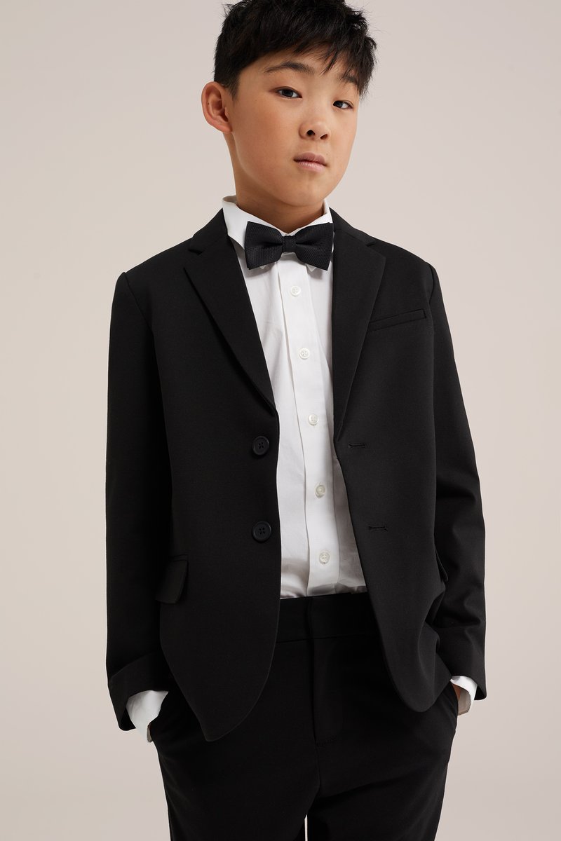 Ragazzo giovane che indossa un completo nero, camicia bianca e papillon nero, in piedi con le mani in tasca davanti a uno sfondo semplice.