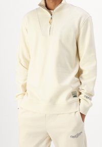 Felpa color crema con collo a mezza zip, dettagli a coste e logo sulla manica. Tessuto morbido con vestibilità comoda.