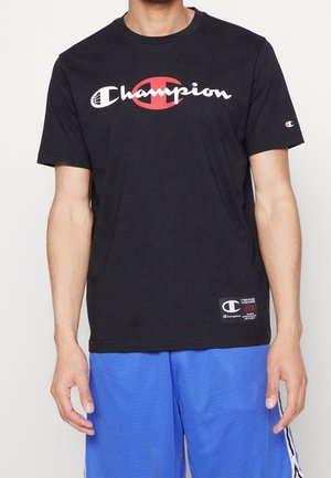 Homme portant un t-shirt noir Champion avec un grand logo et un short en maille bleu, debout devant un fond uni.