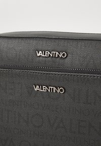 Svart teksturert stoffveske med doble sølv Valentino-logoer. Låsen øverst er en glidelås med subtile sømdetaljer.