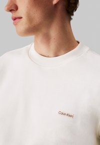Sudadera blanca de cuello redondo hecha de tela texturada. Presenta un pequeño parche con el logotipo rojo de Calvin Klein en la parte izquierda del pecho.