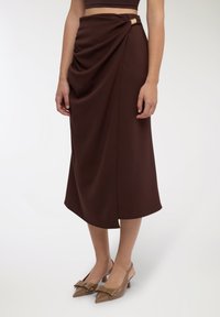 Gonna midi marrone drappeggiata con dettaglio annodato laterale e accento dorato. Indossata con décolleté beige a punta con un fiocco decorativo.