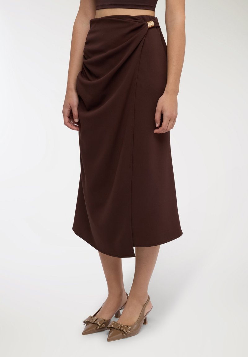 Gonna midi marrone drappeggiata con dettaglio annodato laterale e accento dorato. Indossata con décolleté beige a punta con un fiocco decorativo.