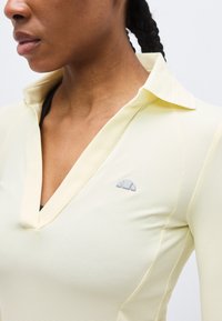 Γυναίκα που φοράει ένα ανοιχτό κίτρινο μακρυμάνικο πουκάμισο με υφή, γιακά και μικρό γκρι λογότυπο στο στήθος, με μαύρα πλεκτά μαλλιά μαζεμένα προς τα πίσω.