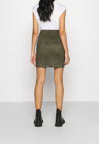 Olivgrön, suéde mini kjol med en figurnära design och en dragkedja bak, tillsammans med svarta ankelstövlar och en vit t-shirt.