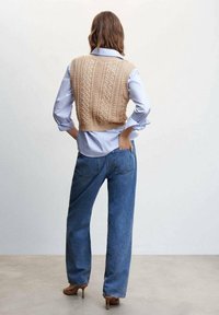 Beige gestrickte, kurze Weste mit Kabelmuster, getragen über einem blauen Hemd mit Knöpfen, kombiniert mit hochtaillierten blauen Jeans und braunen Absätzen.