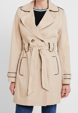 Gabardina beige cruzada con doble botonadura, con ribete negro en los bolsillos y puños, cintura con cinturón, llevada sobre una camiseta negra y vaqueros azules.