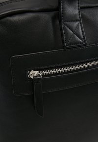 Section de sac en cuir noir avec une poche zippée horizontale en argent et une sangle de poignée cousue au-dessus.