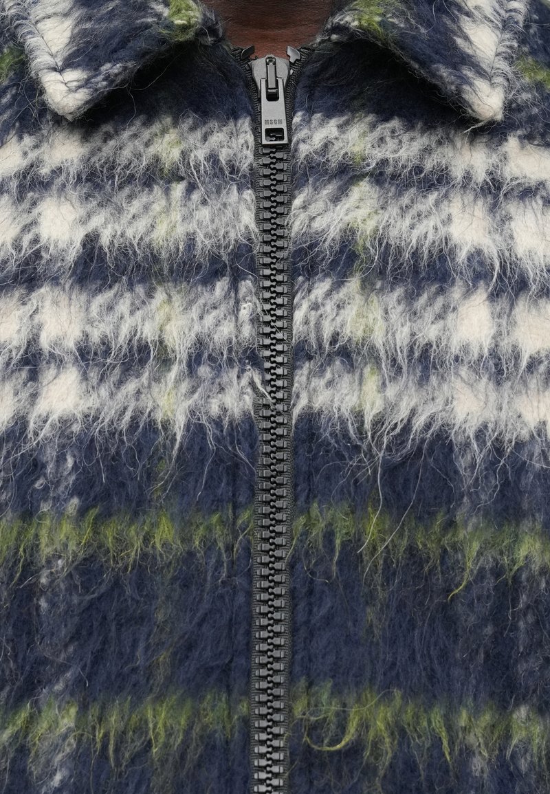 Primer plano de una chaqueta de cuadros suave en azul, blanco y verde, con una cremallera negra y un cuello.