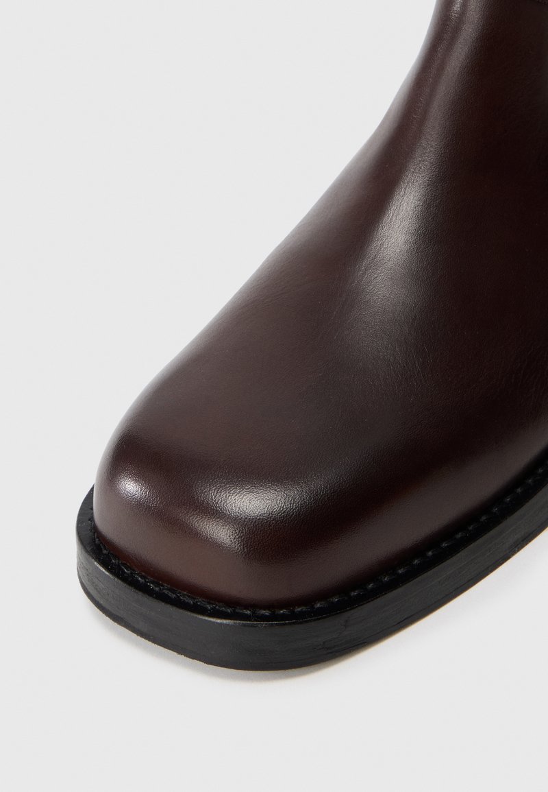 Bottine en cuir marron avec un bout arrondi, texture lisse, semelle noire contrastante et détails de couture le long du bord.