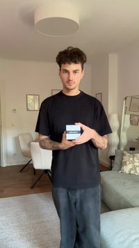 En svart t-skjorte og mørke jeans bært av en person som holder en kvadratisk, lyseblå boks merket "Calvin Klein" foran et minimalistisk oppholdsrom.