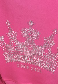 Różowa bluza z kapturem z wzorem korony wykonanym z dżetów, z napisem "OD 2002" poniżej dekoracyjnego wzoru.