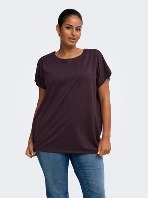 Kvinde iført en mørkelilla kortærmet skjorte og blå jeans, stående mod en ensfarvet lys baggrund.