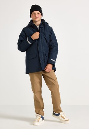 Teenager iført sort hue, navyblå vinterjakke med reflekterende manchetter, beige bukser og hvide sneakers, stående mod ensfarvet baggrund.