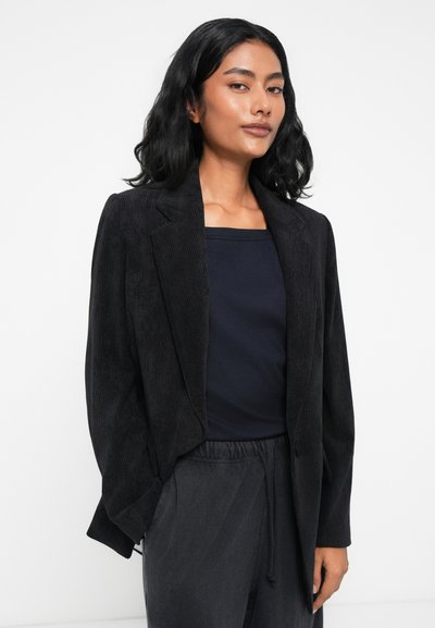 Blazer en velours côtelé noir avec revers cranté, fermeture à un bouton, et coupe décontractée, associé à un haut bleu marine à encolure carrée et à un pantalon sombre.