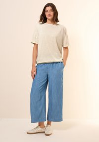 Maglietta grigia chiara a maniche corte con dettagli a macchie, abbinata a pantaloni larghi blu chiaro a gamba larga e sneakers bianche casual.