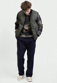 Prošívaná zelená bomber bunda přes vícebarevný kostkovaný svetr, doplněná tmavě modrými kalhotami a béžovými teniskami, stojící postoj.